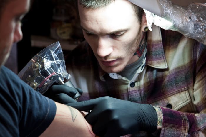 Evil From the Needle ist Camden Towns erstes und am längsten laufendes Tattoo-Studio. Es ist nicht nur eines der Top 5 Londons, sondern wurde ursprünglich 1986 vom weltberühmten Tätowierer Bugs eröffnet. Es ist jetzt im Besitz des angesehenen New Yorker Tätowierers Jeff Ortega. Dave Bryant, ein bekannter Tätowierer bei Evil From the Needle, skizziert und schattiert ein Tattoo auf Chris King.