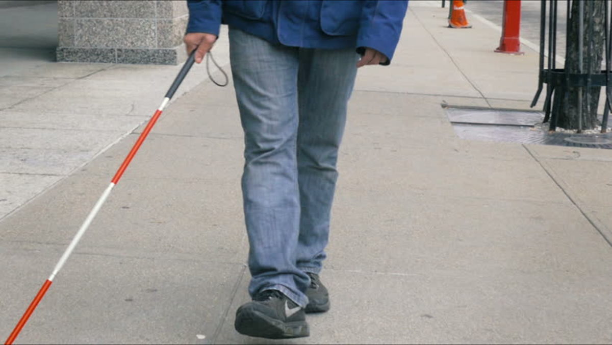 Fotobeschreibung: Mann in Jeans und Turnschuhen, der mit Gehstock auf Bürgersteig geht